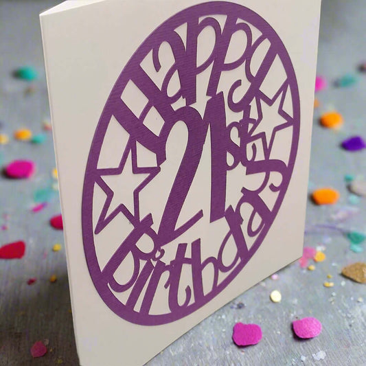 Angled view of a purple papercut 'Happy 21st Birthday' card with stars, displayed on a glittery background scattered with colourful confetti.