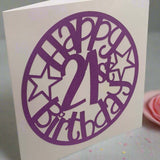 Angled view of a purple 'Happy 21st Birthday' papercut card with star accents, displayed on a soft pastel background with confetti and a pink rosette.