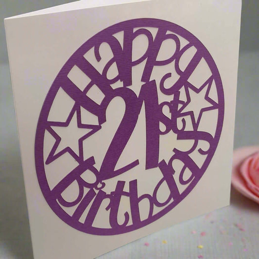 Angled view of a purple 'Happy 21st Birthday' papercut card with star accents, displayed on a soft pastel background with confetti and a pink rosette.