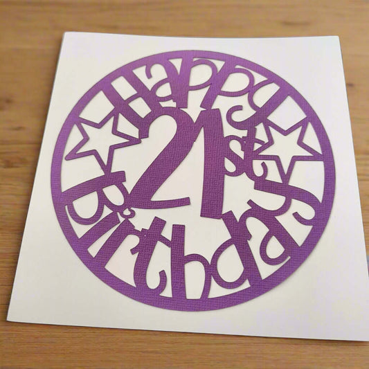 Purple papercut topper reading 'Happy 21st Birthday' in a circle design with stars, displayed on white card against a wooden background.