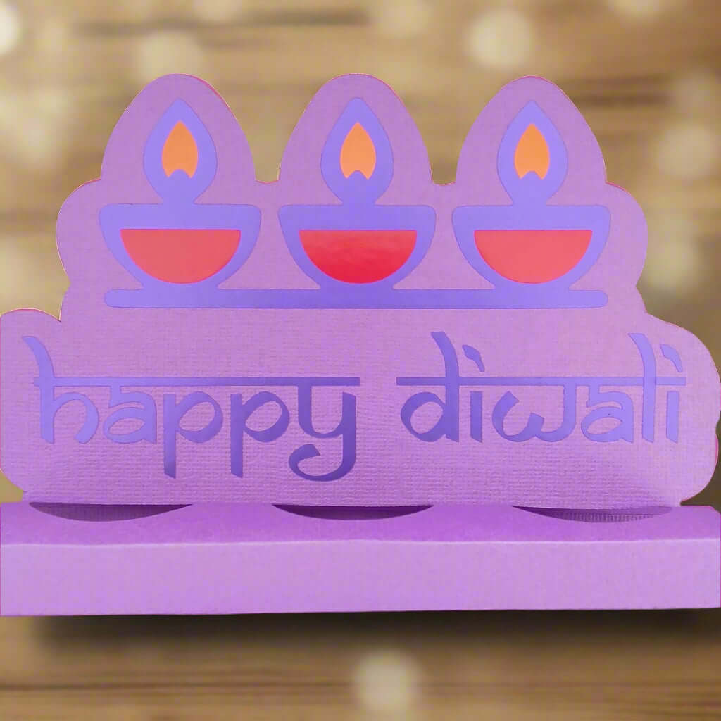 Straight-on image of a purple Happy Diwali sweet holder made from textured cardstock with three layered diya designs, set against a festive blurred golden bokeh background.