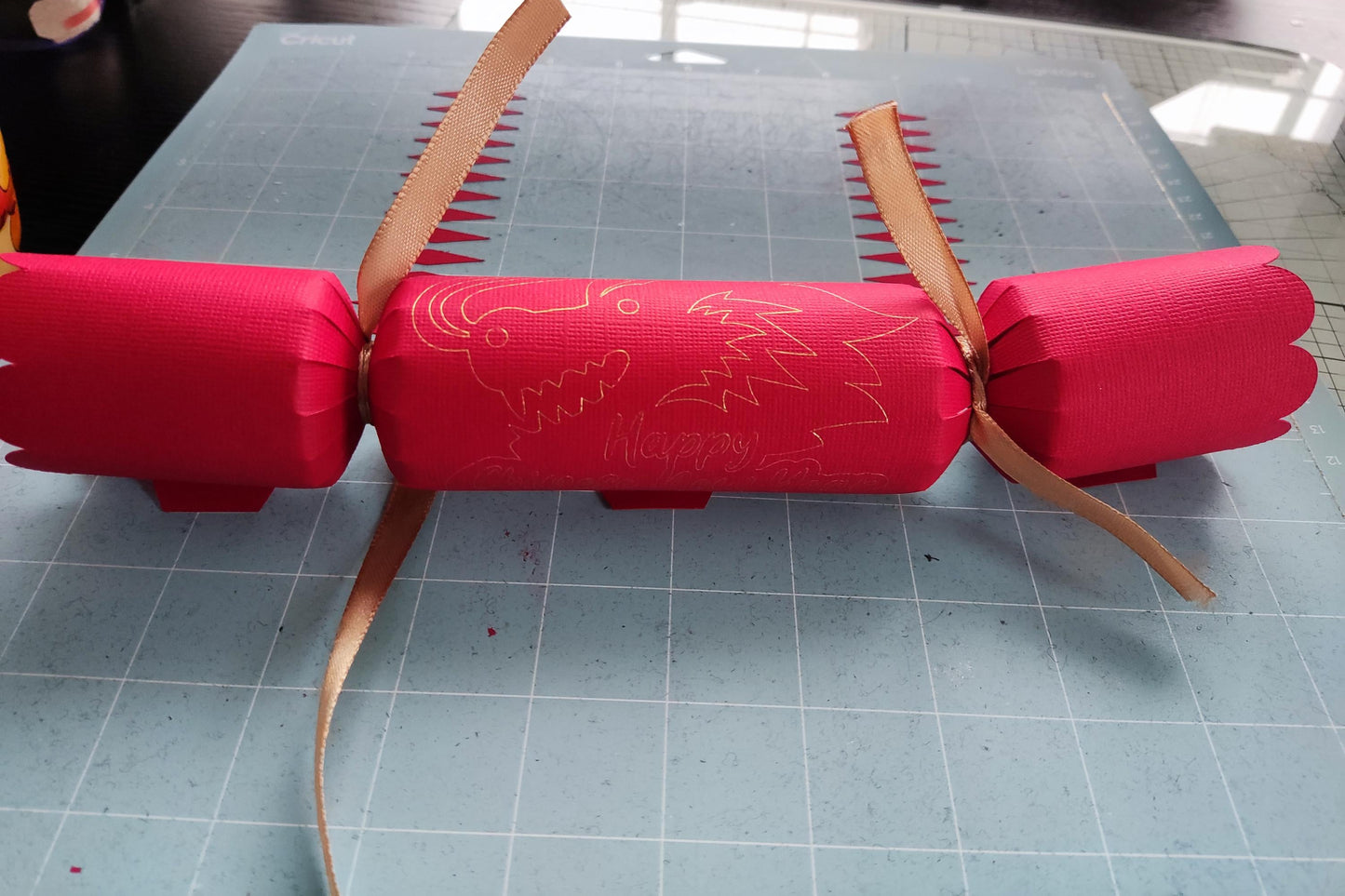 Handmade red Chinese New Year cracker with gold foil dragon design, tied with gold ribbon, shown flat on a crafting mat during assembly.