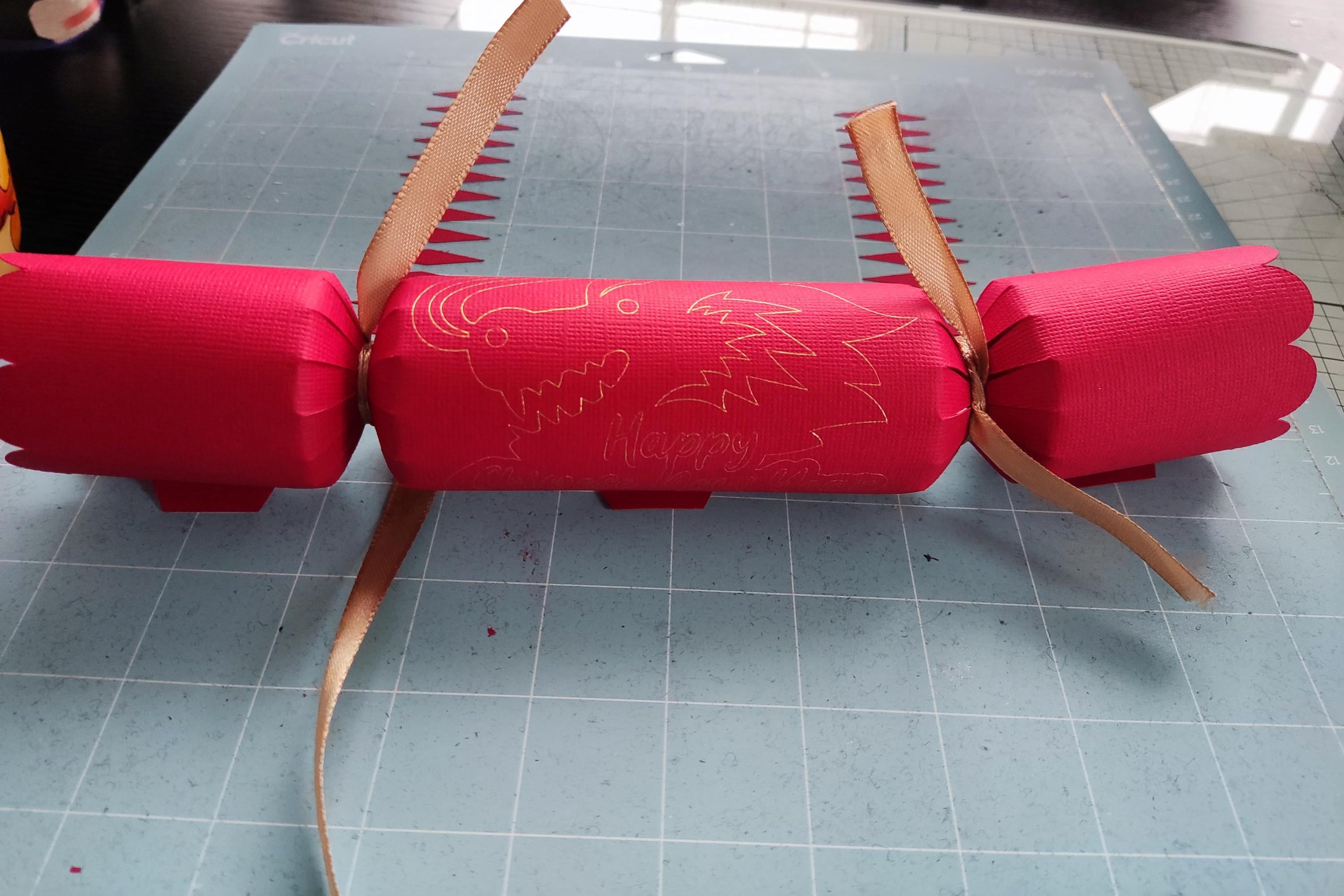Handmade red Chinese New Year cracker with gold foil dragon design, tied with gold ribbon, shown flat on a crafting mat during assembly.