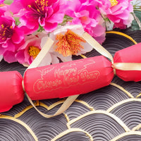 Red Chinese New Year cracker with gold foil “Happy Chinese New Year” design, tied with gold satin ribbon, styled with bright pink blossom flowers on a patterned black and gold background.