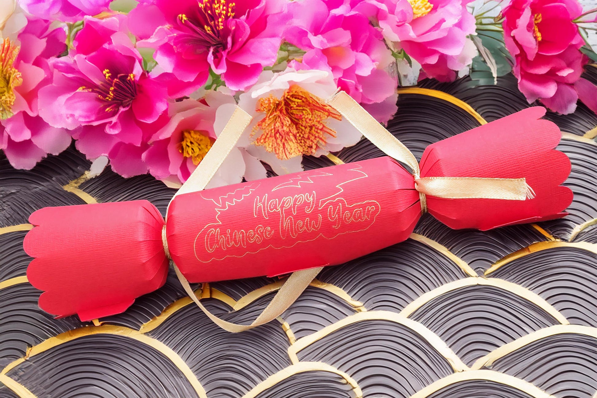 Red Chinese New Year cracker with gold foil “Happy Chinese New Year” design, tied with gold satin ribbon, styled with bright pink blossom flowers on a patterned black and gold background.