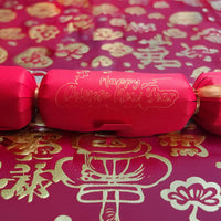 Red cracker with gold foil “Happy Chinese New Year” lettering, tied with gold ribbon, displayed on a red and gold patterned festive backdrop with traditional Chinese motifs.