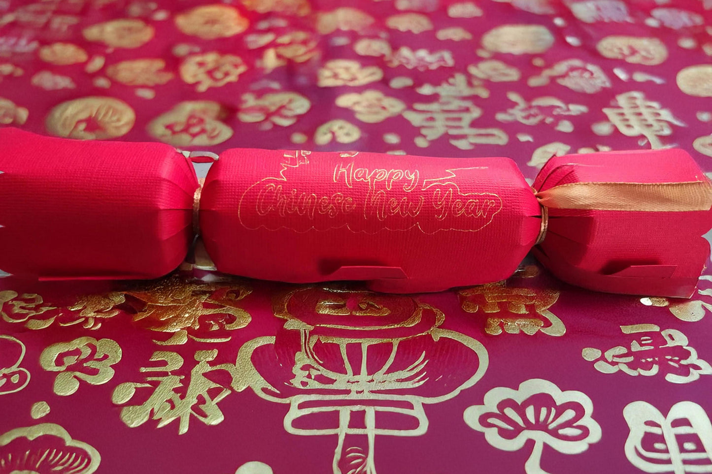 Red cracker with gold foil “Happy Chinese New Year” lettering, tied with gold ribbon, displayed on a red and gold patterned festive backdrop with traditional Chinese motifs.