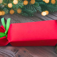 Plain red cracker tied with green ribbon, displayed on rustic wooden table with pinecones and festive garland with warm golden lights.