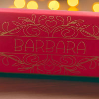 Close-up of red cracker with intricate gold foil design and name “Barbara,” captured with warm bokeh lights in the background for a festive glow.