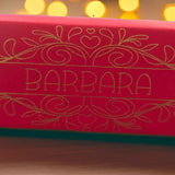 Close-up of red cracker with intricate gold foil design and name “Barbara,” captured with warm bokeh lights in the background for a festive glow.