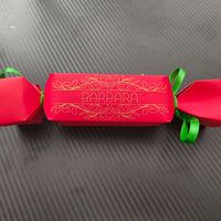 Red cracker with gold foil design and name “Barbara,” tied with green ribbon, photographed flat on textured black background for modern contrast.