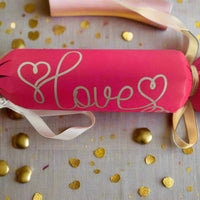 Close-up of a red cracker with “Love” in gold pen, tied with ribbon and surrounded by golden heart confetti, perfect for weddings or Valentine’s.
