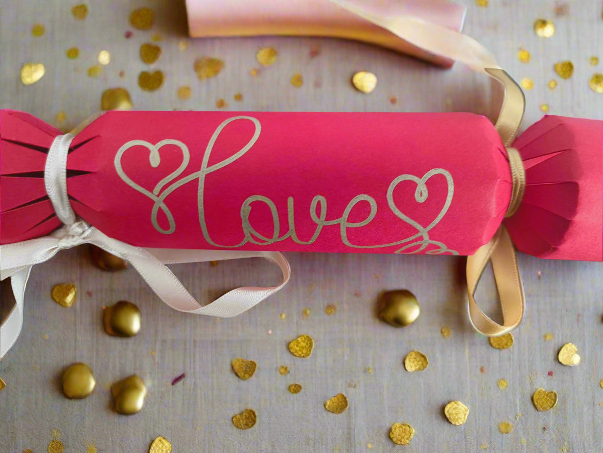 Close-up of a red cracker with “Love” in gold pen, tied with ribbon and surrounded by golden heart confetti, perfect for weddings or Valentine’s.

