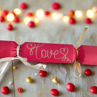 Red cracker with “Love” hand-lettered in gold pen, tied with ivory ribbon, styled with glowing lights and red and gold beads for a festive effect.
