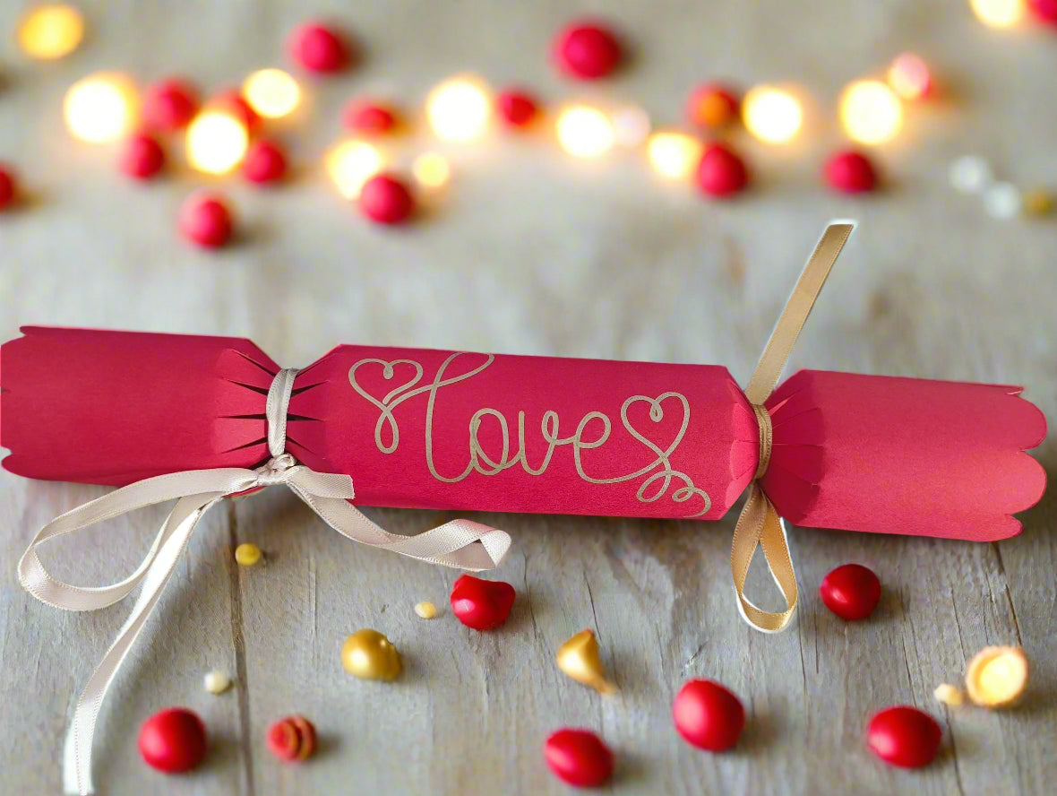 Red cracker with “Love” hand-lettered in gold pen, tied with ivory ribbon, styled with glowing lights and red and gold beads for a festive effect.