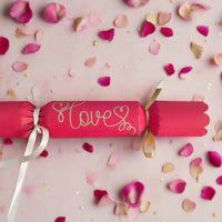 Red cracker with “Love” written in gold pen, tied with white and gold ribbons, surrounded by scattered pink rose petals for a romantic theme.