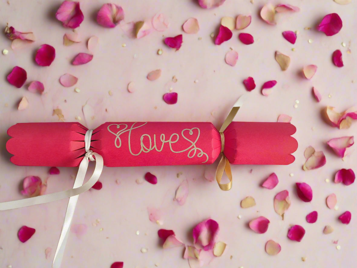Red cracker with “Love” written in gold pen, tied with white and gold ribbons, surrounded by scattered pink rose petals for a romantic theme.