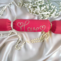 A red cracker with “Love” hand-drawn in gold pen, tied with white ribbon, styled on satin with pearls and white flowers for a romantic look.
