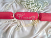 A red cracker with “Love” hand-drawn in gold pen, tied with white ribbon, styled on satin with pearls and white flowers for a romantic look.