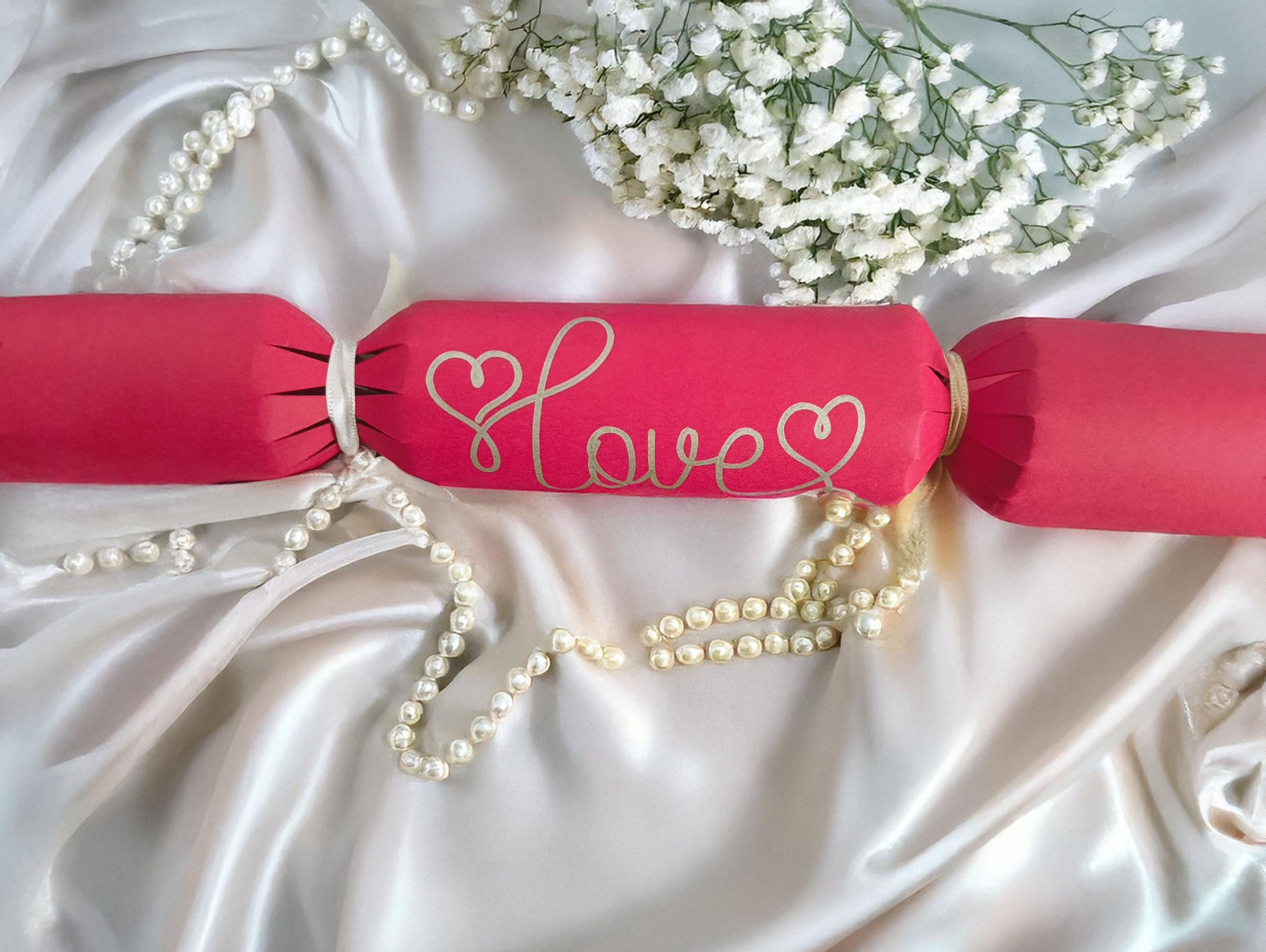 A red cracker with “Love” hand-drawn in gold pen, tied with white ribbon, styled on satin with pearls and white flowers for a romantic look.