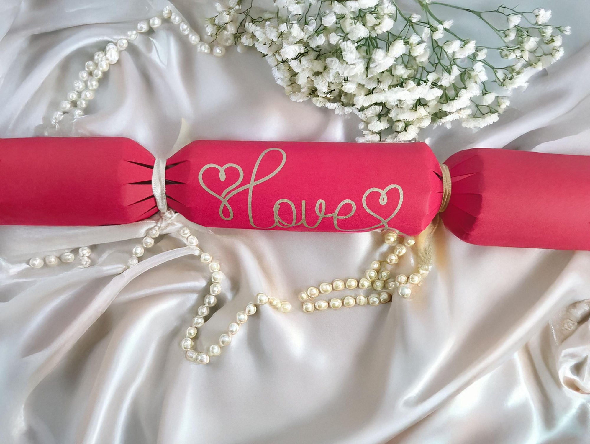 A red cracker with “Love” hand-drawn in gold pen, tied with white ribbon, styled on satin with pearls and white flowers for a romantic look.