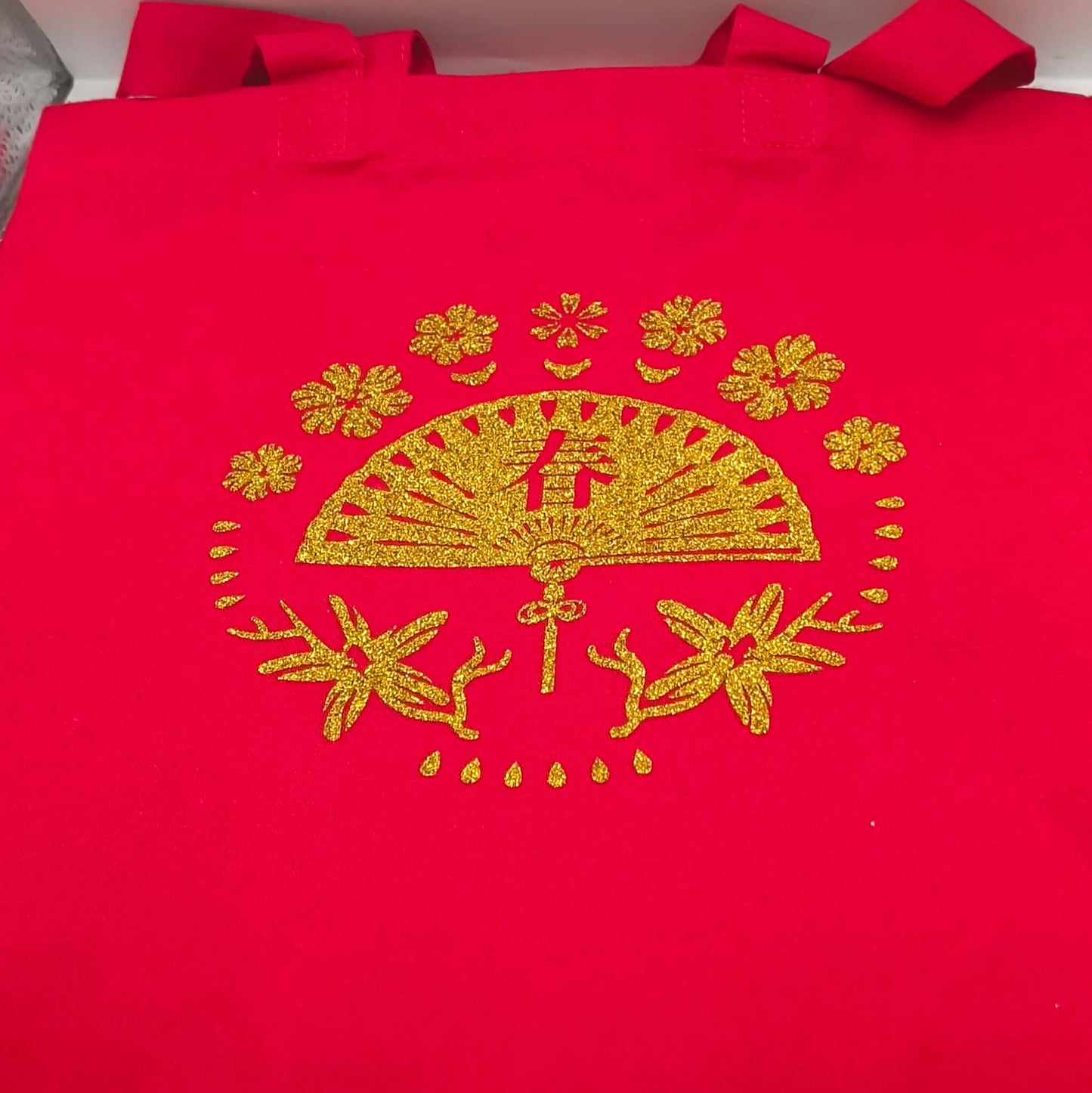 Full view of a red tote bag decorated with a gold glitter fan and floral design, displayed standing upright.