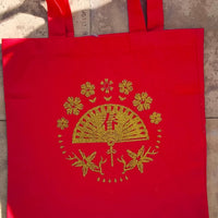 Back view of red tote bag with gold glitter fan and floral vinyl design, laid flat on stone background.