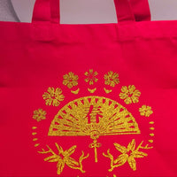 Red fabric tote bag with sturdy handles, featuring a glitter gold fan and floral design at the centre.
