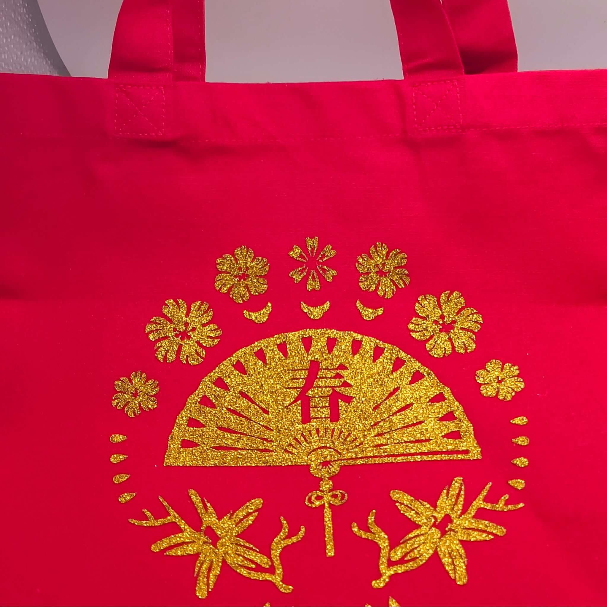 Red fabric tote bag with sturdy handles, featuring a glitter gold fan and floral design at the centre.