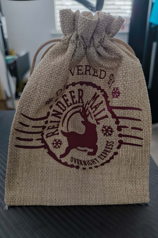 Standing hessian gift bag tied with drawstring, featuring "Reindeer Mail Overnight Express" festive burgundy design.