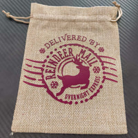 Hessian Christmas gift bag with burgundy "Delivered by Reindeer Mail Overnight Express" design, flat and empty on black background.