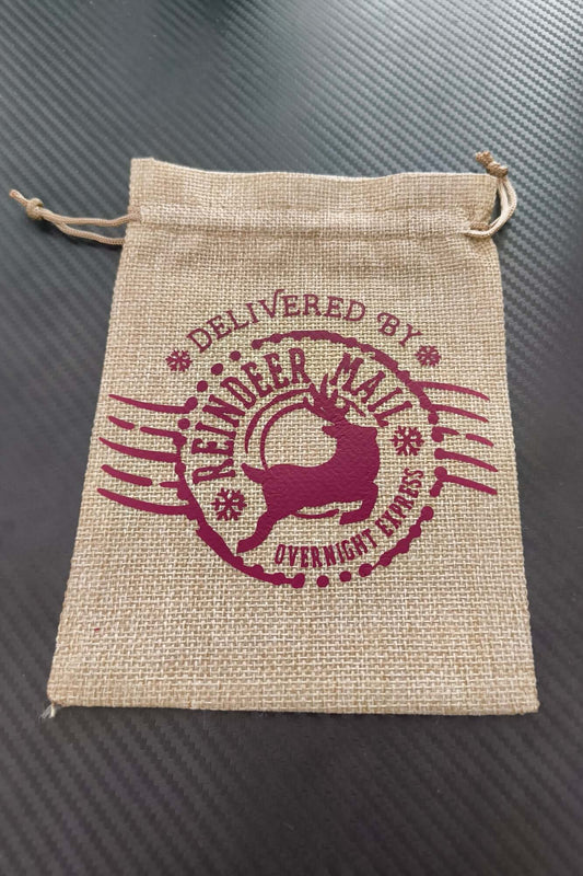 Hessian Christmas gift bag with burgundy "Delivered by Reindeer Mail Overnight Express" design, flat and empty on black background.