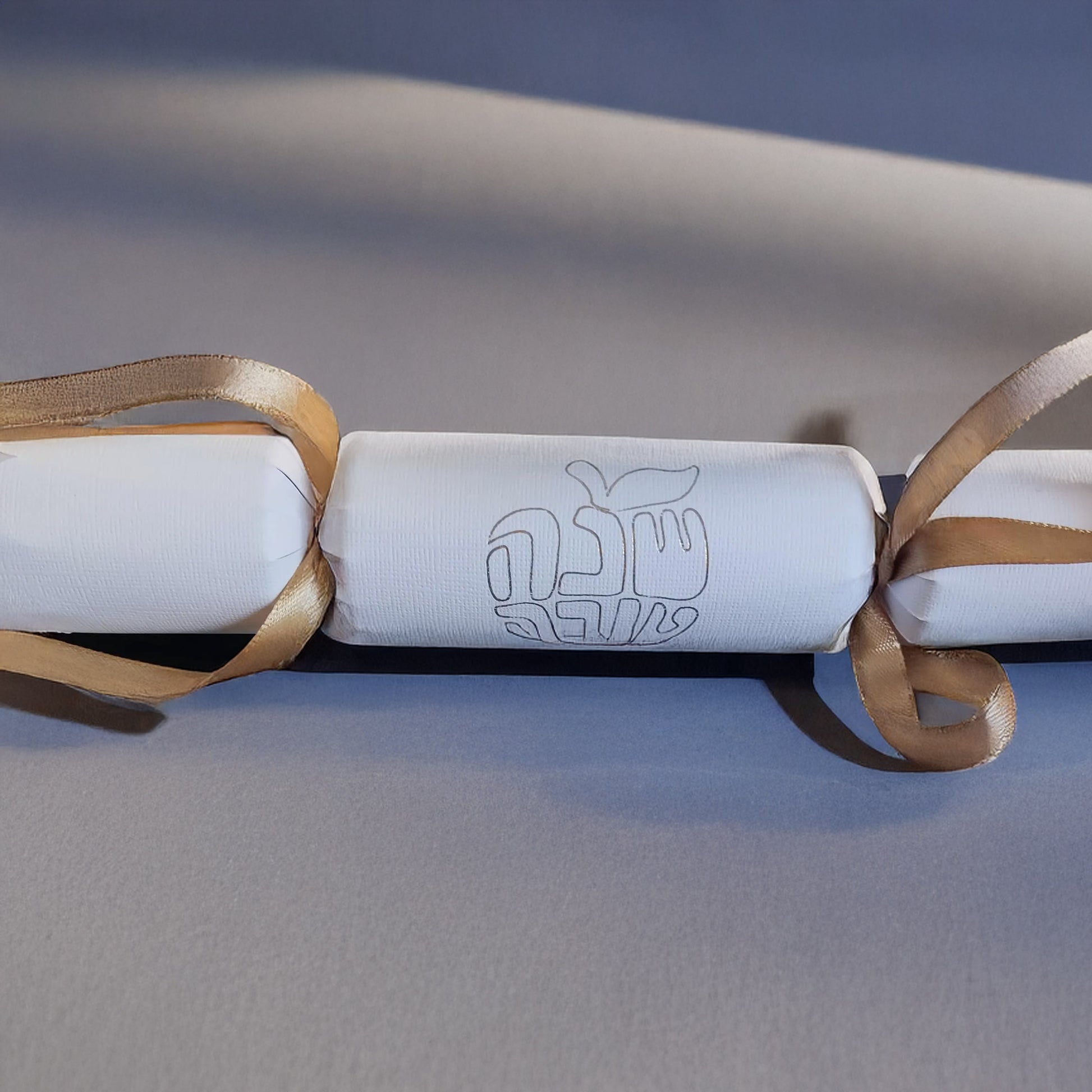 White cracker with gold foil Hebrew “Shana Tova” design and gold ribbon, displayed on a neutral background.
