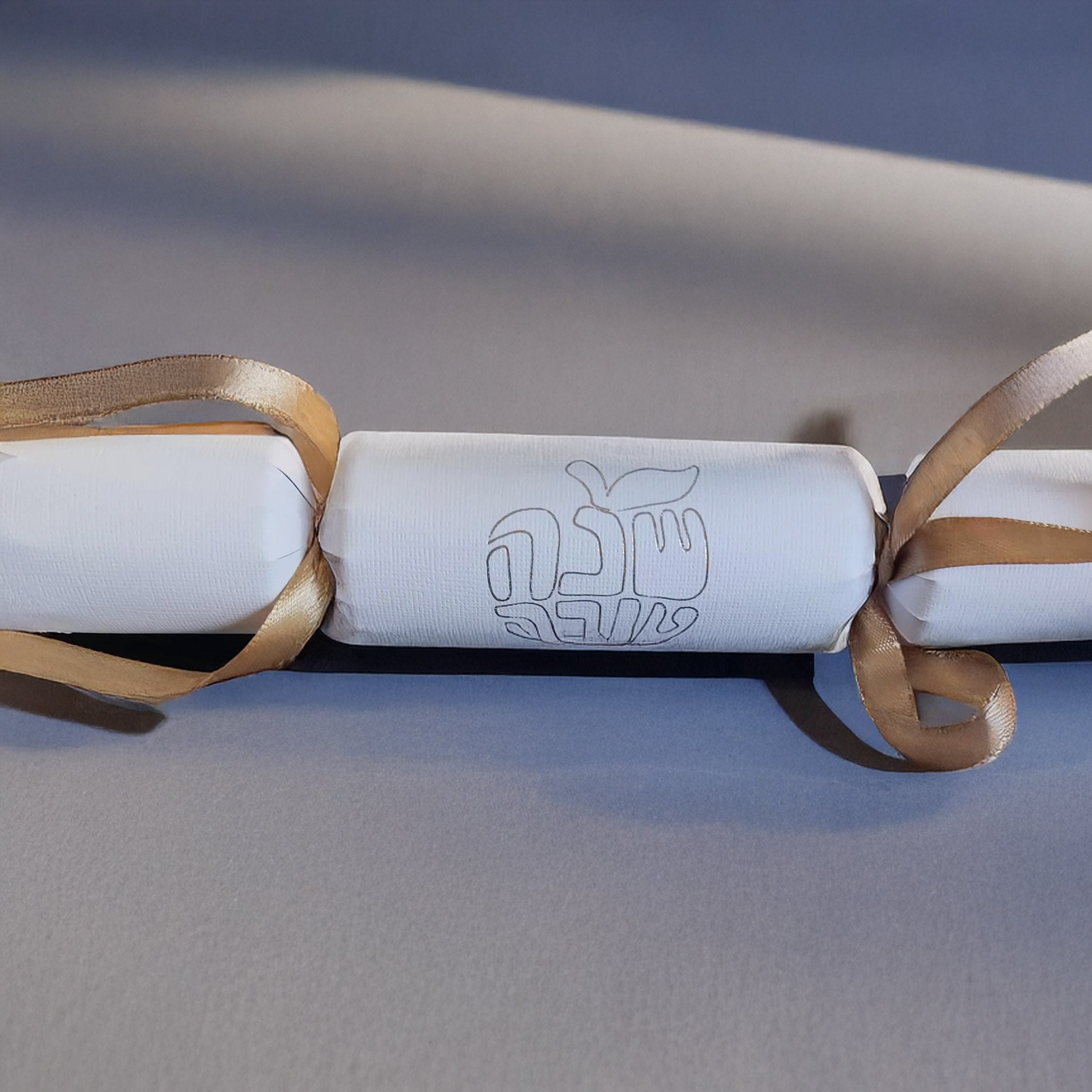White cracker with gold foil Hebrew “Shana Tova” design and gold ribbon, displayed on a neutral background.
