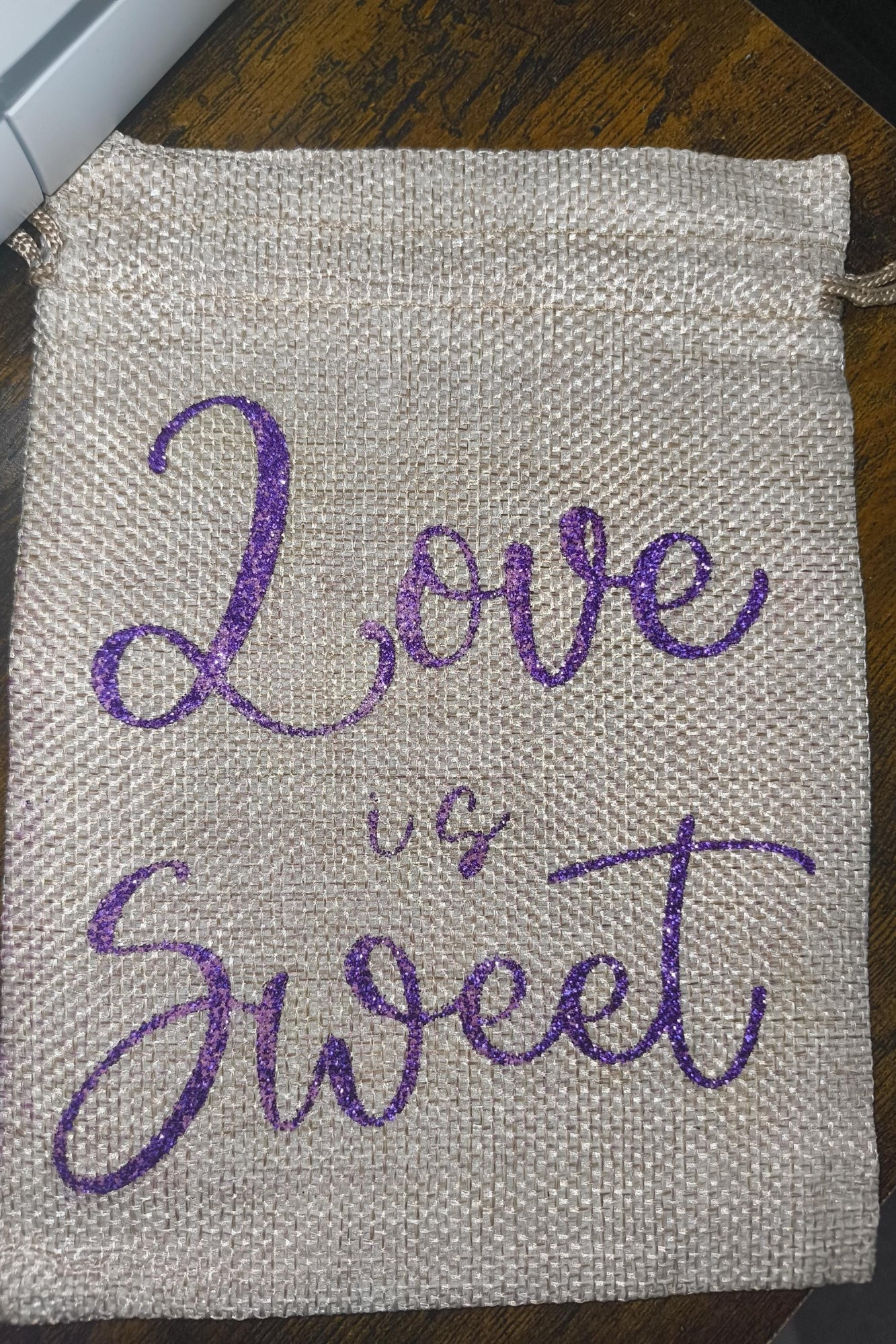 Close-up of a hessian favour bag with shimmering purple glitter lettering “Love is Sweet.” Woven, textured finish with drawstring closure, perfect for weddings or gifts.