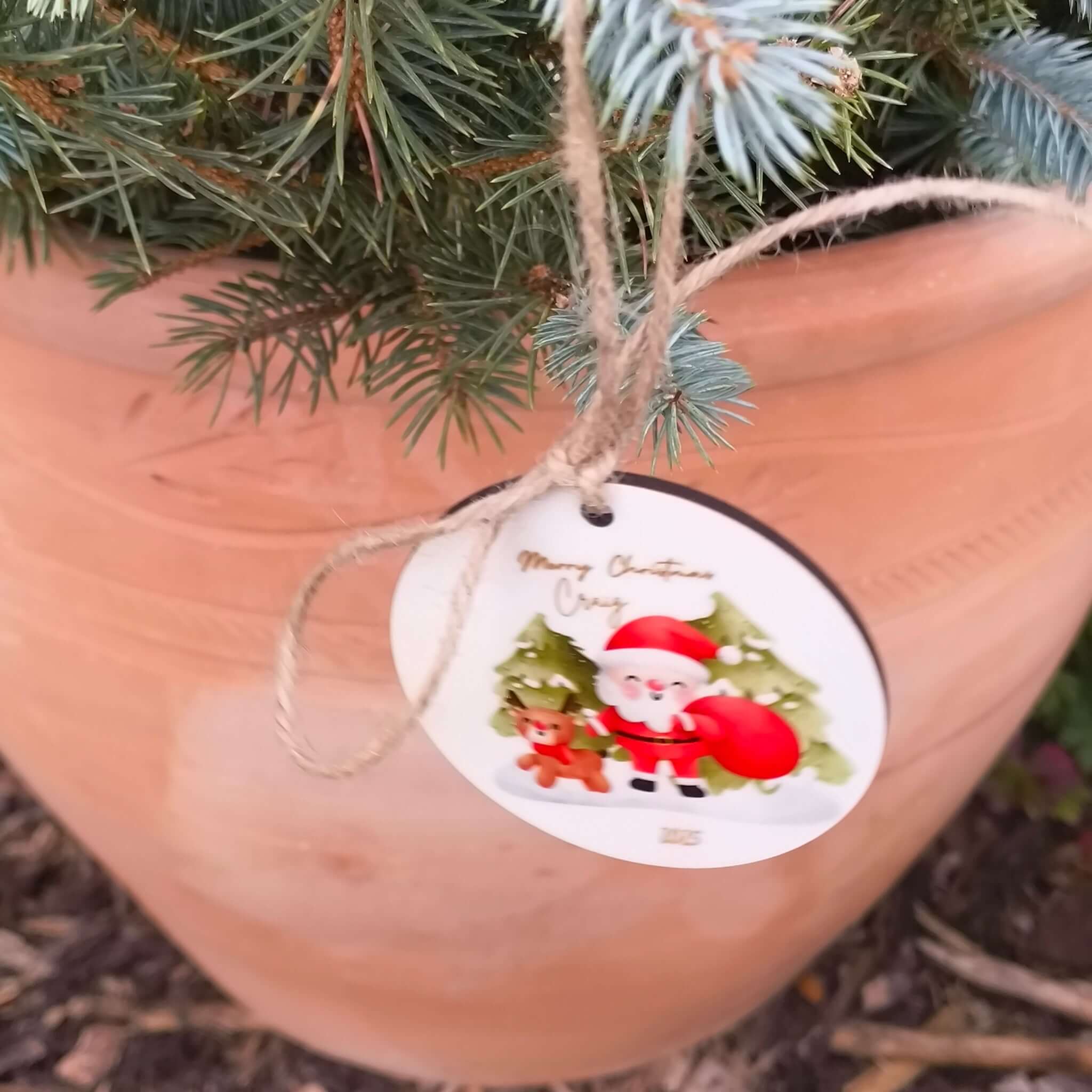 Close-up of Santa Christmas bauble with custom name, tied with jute string on festive evergreen branches.