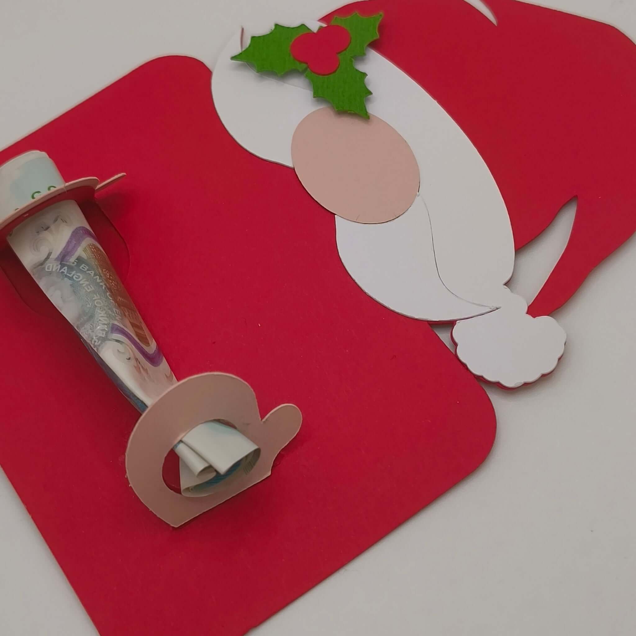 Angled view of a handmade Santa money holder card with hat and holly detail, holding a rolled £5 banknote between two cut-out slots.