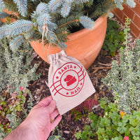 Close-up of Santa Mail hessian drawstring bag with red festive design against pine tree background.