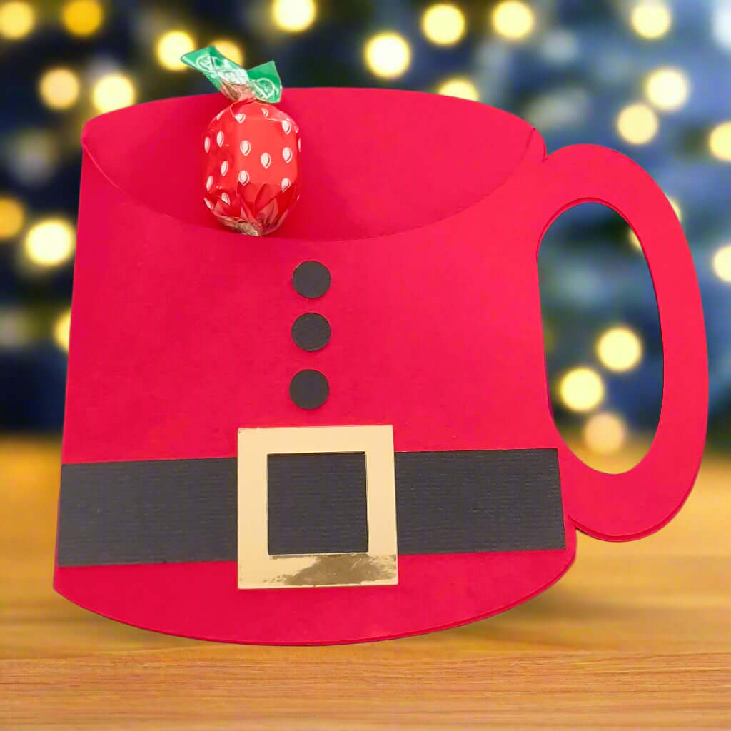 Handcrafted Santa suit mug-shaped gift card holder displayed on a wooden table with blurred Christmas lights in the background – perfect for festive gifting, stocking fillers, or table favours – chocolates not included.