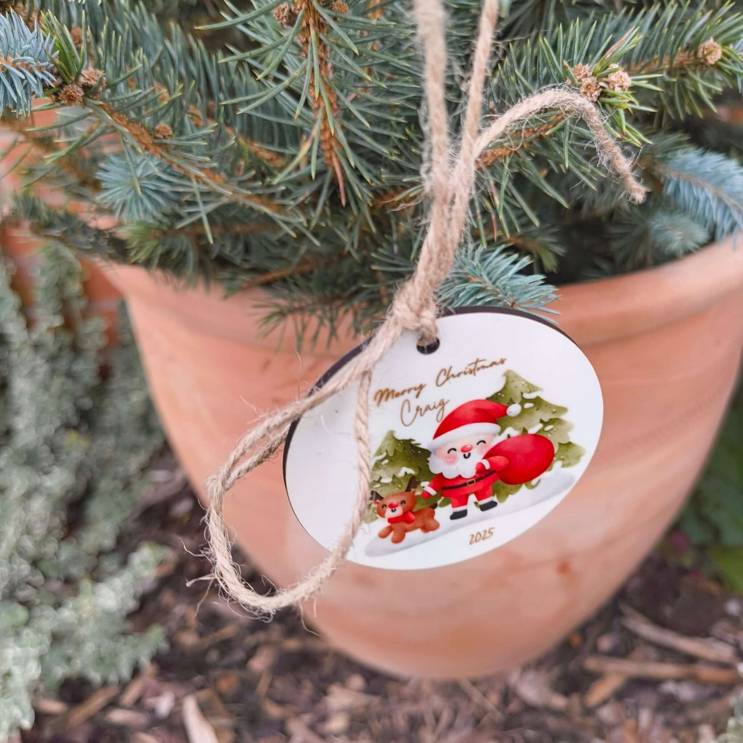 Festive Santa and reindeer Christmas bauble with natural twine, displayed on a potted evergreen tree.