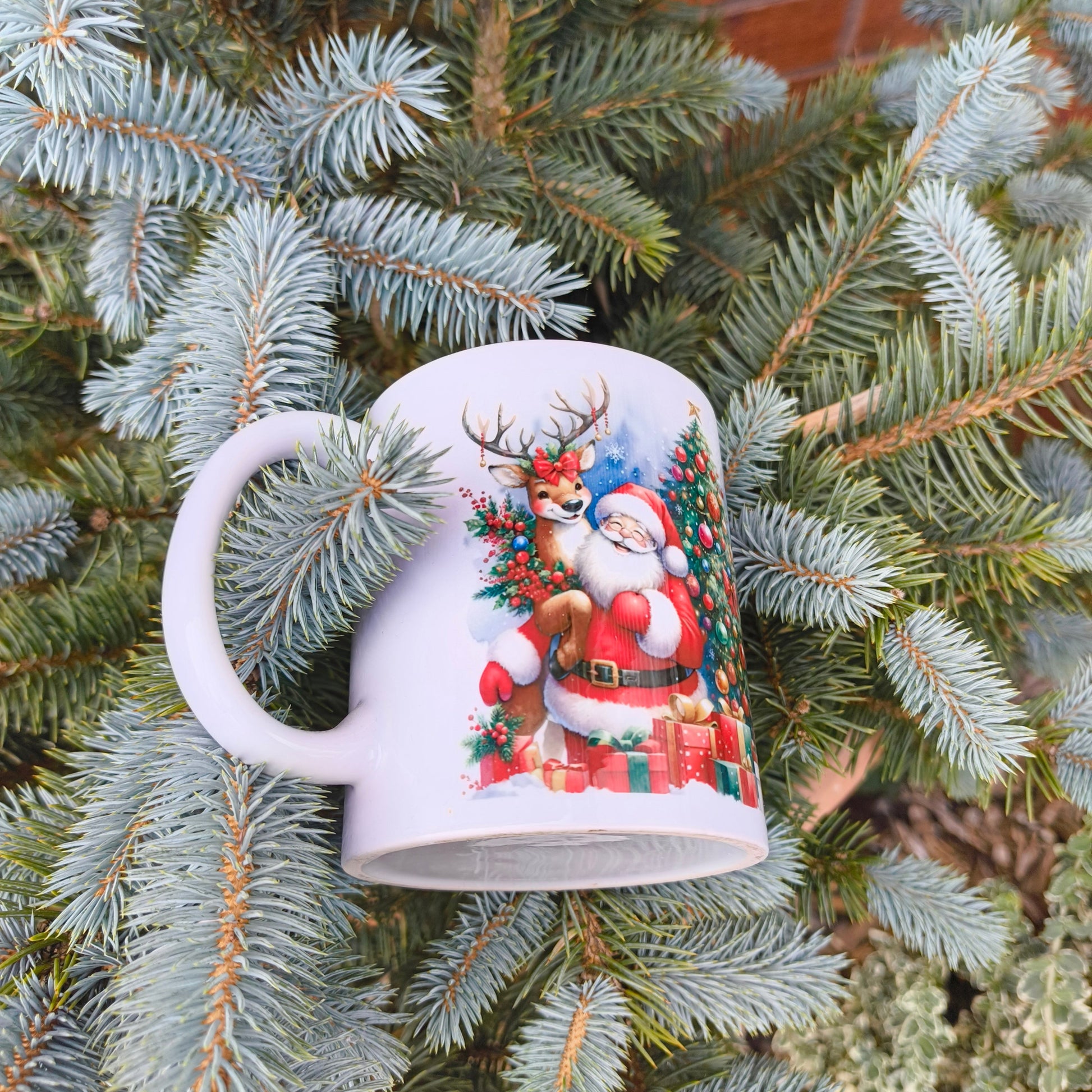 White ceramic Christmas mug with Santa Claus, reindeer, and festive tree design, displayed on a pine branch outdoors