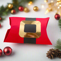 Red Santa-themed pillow gift box with a gold buckle and black belt design, surrounded by festive Christmas decorations including pinecones, red and gold baubles, fir branches, and warm fairy lights on a soft white background.

