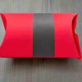 Back view of a handmade red Christmas pillow gift box with a wide black belt design, displayed on a light wood grain background for a clean and festive presentation.

