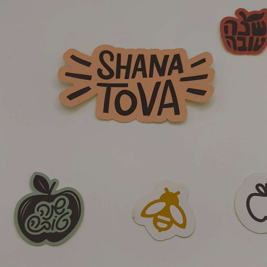 Close-up of Shana Tova stickers showing a bold text design, apple outline, bee illustration, and apple with Hebrew lettering.