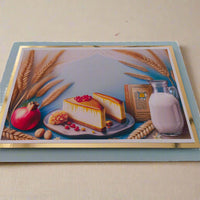 Handmade Shavuot greeting card featuring a cheesecake, milk jug, wheat stalks, pomegranate, and prayer book, displayed on a soft cream fabric background.