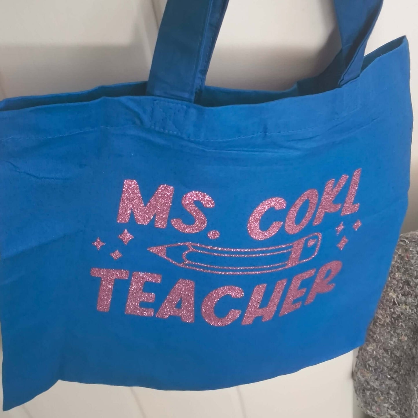 Handmade blue cotton tote bag with pink glitter vinyl text "Ms. Cohl Teacher," hanging neatly on a silver door handle.