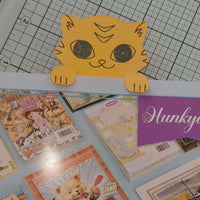 Cute tiger cat paper bookmark peeking over edge, top-down angle on cutting mat background