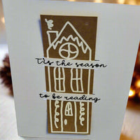 Handmade Christmas card featuring a brown gingerbread-style house design with white icing details. Across the card, black cursive text reads: '’Tis the season to be reading'. The background is softly lit with warm, festive lights.