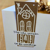 Handmade Christmas card featuring a tall gingerbread-style house design in white on a brown cut-out. The text over the house reads: '’Tis the season to be reading' in black cursive font. The card is photographed in warm lighting with pinecones and festive lights in the background