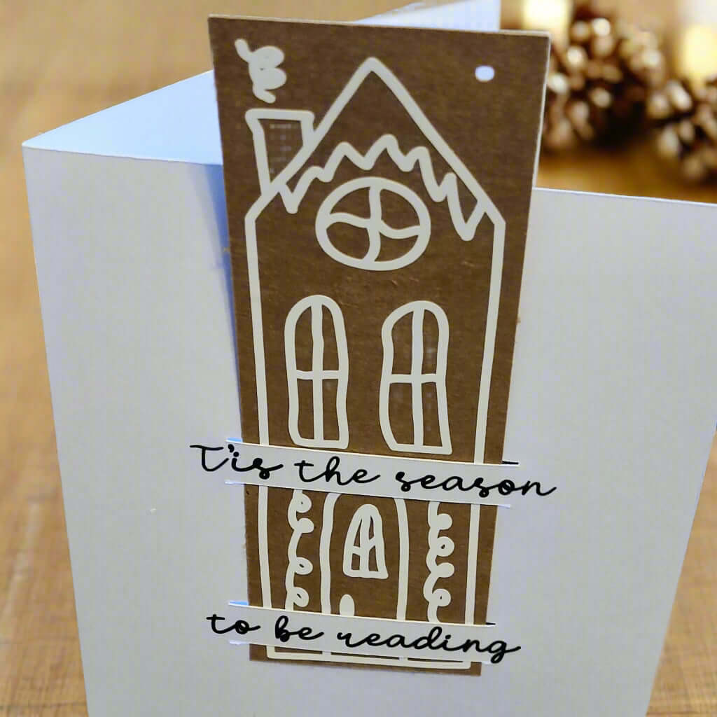 Handmade Christmas card featuring a tall gingerbread-style house design in white on a brown cut-out. The text over the house reads: '’Tis the season to be reading' in black cursive font. The card is photographed in warm lighting with pinecones and festive lights in the background